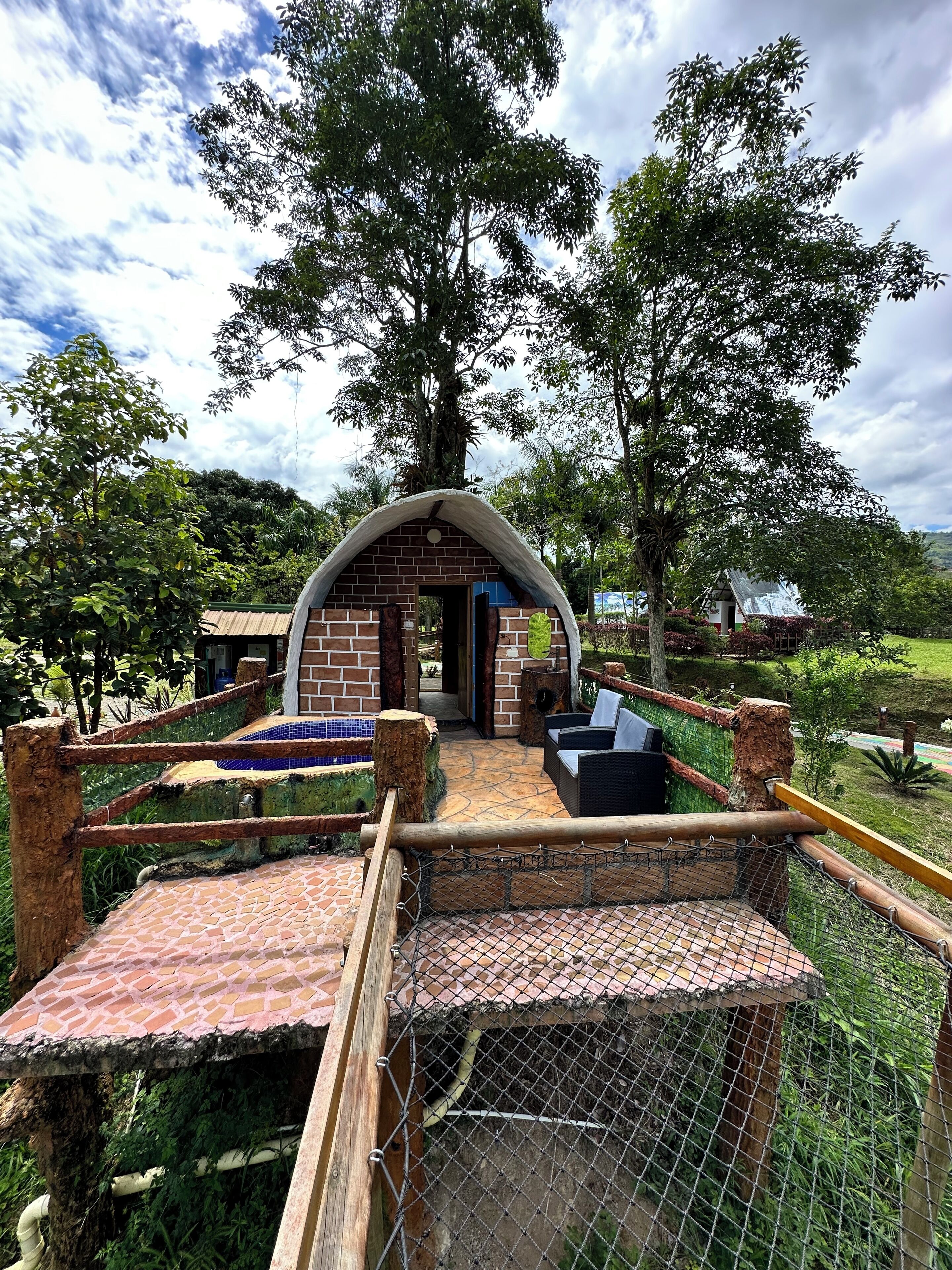 Un espace extérieur aménagé dans un style rustique, avec une arche en briques, entouré d’une végétation luxuriante et sous un ciel dégagé.