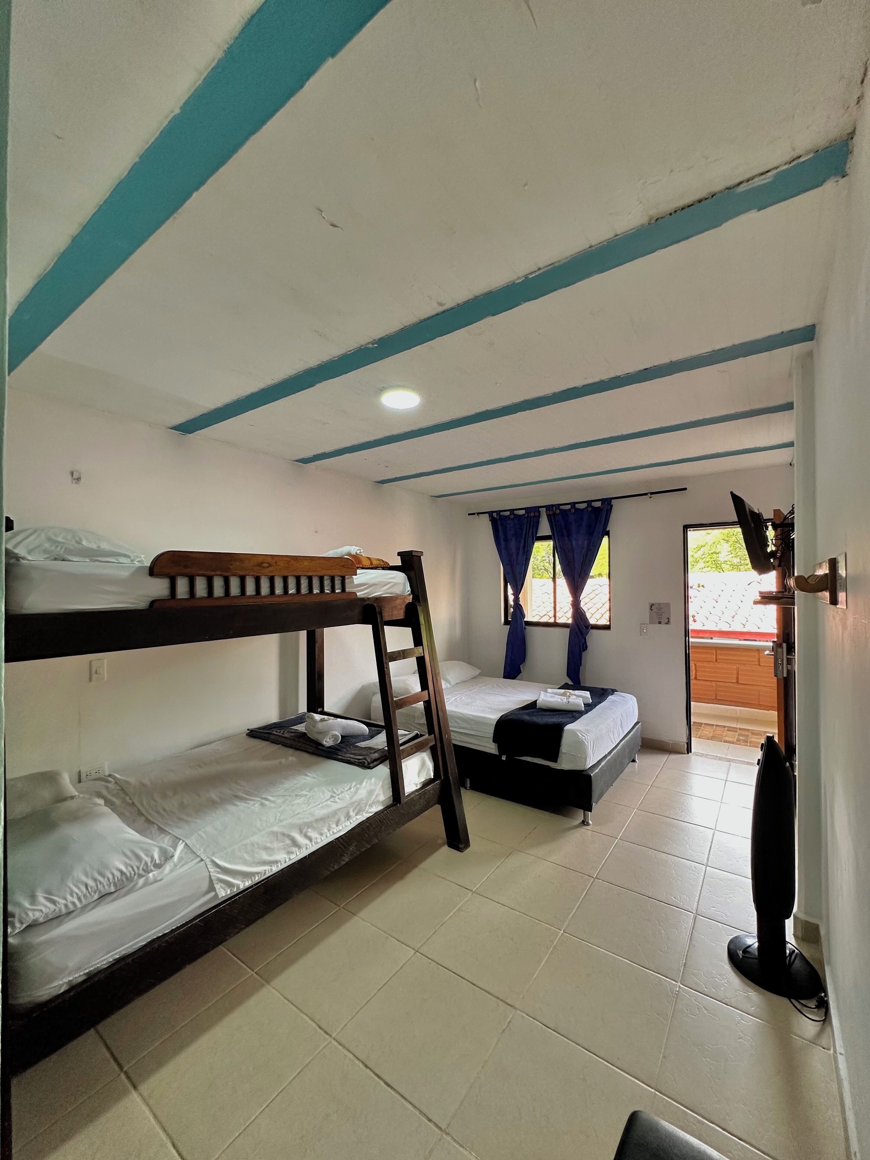 Une chambre de dortoir avec des lits superposés, un lit simple, une fenêtre avec des rideaux, un téléviseur et une porte donnant sur l’extérieur.