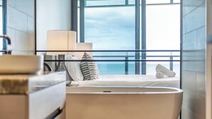 Een moderne hotelkamer met een groot raam, een bed met witte lakens, een nachtkastje met een lamp en uitzicht op zee.