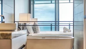 Een moderne hotelkamer met een groot raam, een bed met witte lakens, een nachtkastje met een lamp en uitzicht op zee.