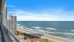 Een hoog gebouw met uitzicht op een strand, oceaan golven, een heldere lucht en een verre blik op een ander gebouw.