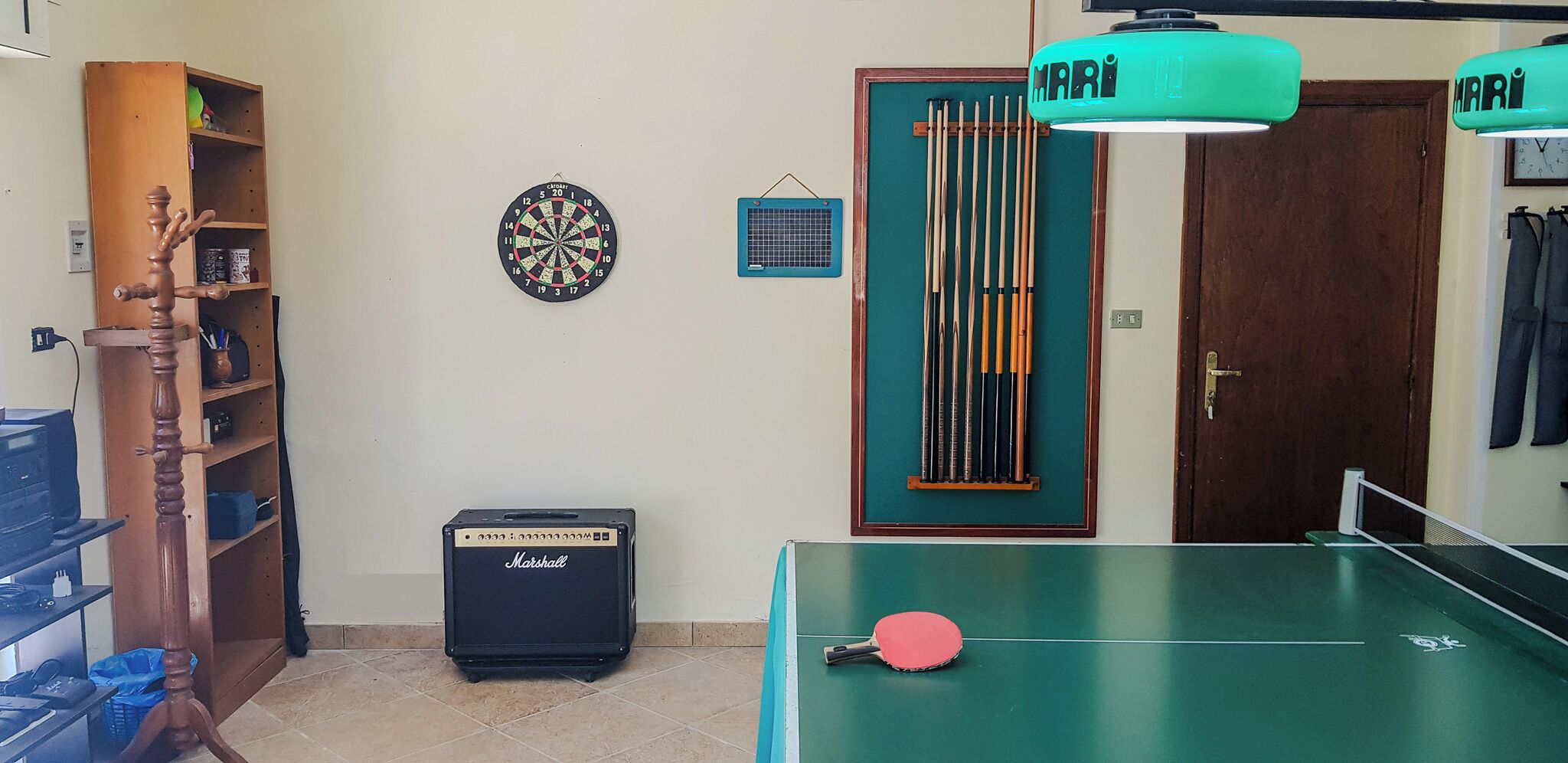 Una habitación con dardos, una mesa de billar, un amplificador Marshall y una puerta con el cartel 'MARI'.