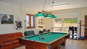 Uma mesa de bilhar com superfície de feltro verde, um sofá de couro marrom e um conjunto de bateria ao fundo.