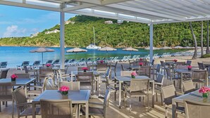 Restaurante frente a la playa con terraza, con vistas a un mar tranquilo y una colina con edificios.