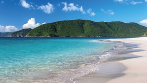 Una playa con agua turquesa cristalina, arena blanca y una colina boscosa al fondo.