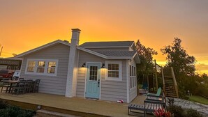 Une petite maison avec une porte bleu clair, une cheminée et une terrasse. Un escalier mène à une zone de terrasse en bois où se trouvent un banc et une chaise.