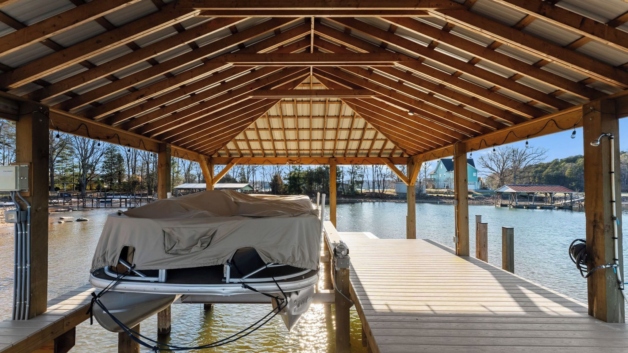 A covered dock with a boat covered in a tarp, wooden beams, and a view of a lake with buildings in the background.