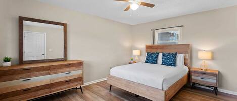 A bedroom with a wooden bed, a nightstand with a lamp, a ceiling fan, a large mirror, and a window with blinds.