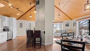 A spacious kitchen with wooden flooring and a high ceiling with exposed beams. There is a dining area with a table set for four, and a view of the outside through large windows. - Pleasant Cove Waterfront Getaway With Fire Pit, Deck, and Lake Views (Mooresville)