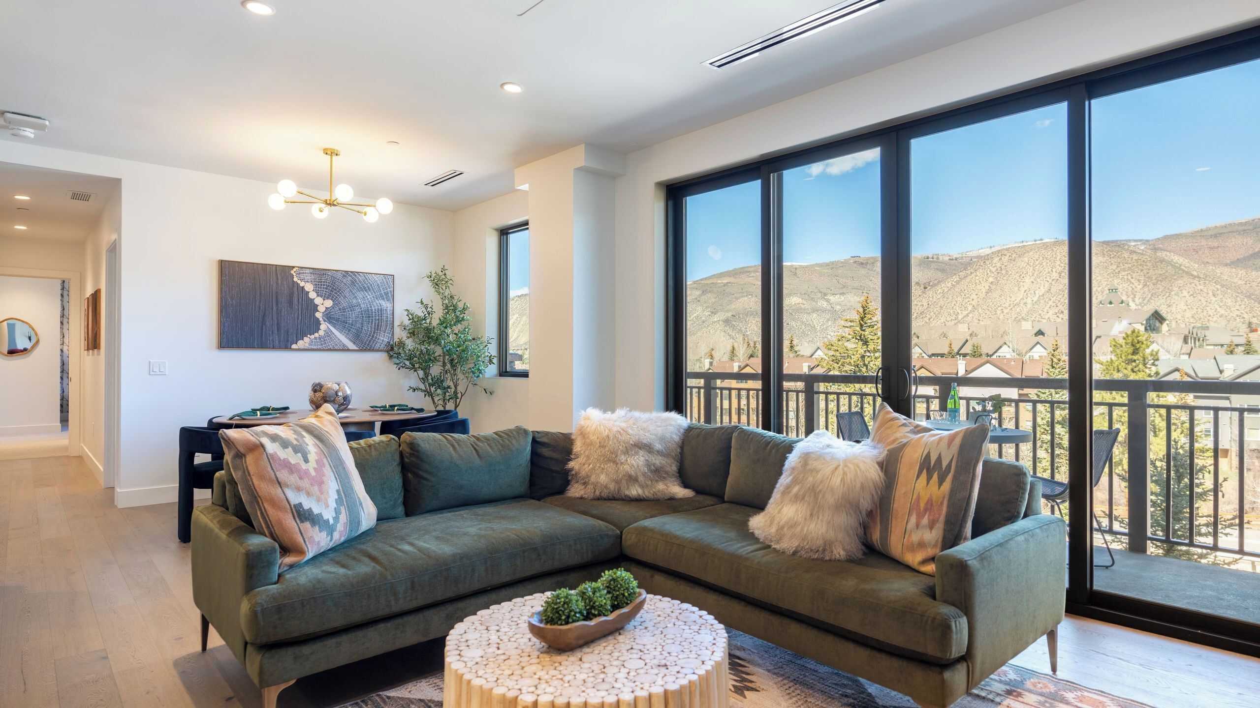 A modern living room with a sectional sofa, a round coffee table with plants, a dining area with a table and chairs, and large windows offering a mountain view.