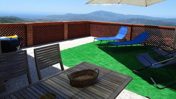 Una terraza con vistas a montañas distantes, una mesa de madera, sillones reclinables azules y un gran paraguas.