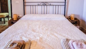 Una cama bien tendida con una colcha estampada, dos mesitas de noche con velas, y una silla al fondo.
