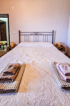 A neatly made bed with a patterned bedspread, two side tables with candles, and a chair in the background.