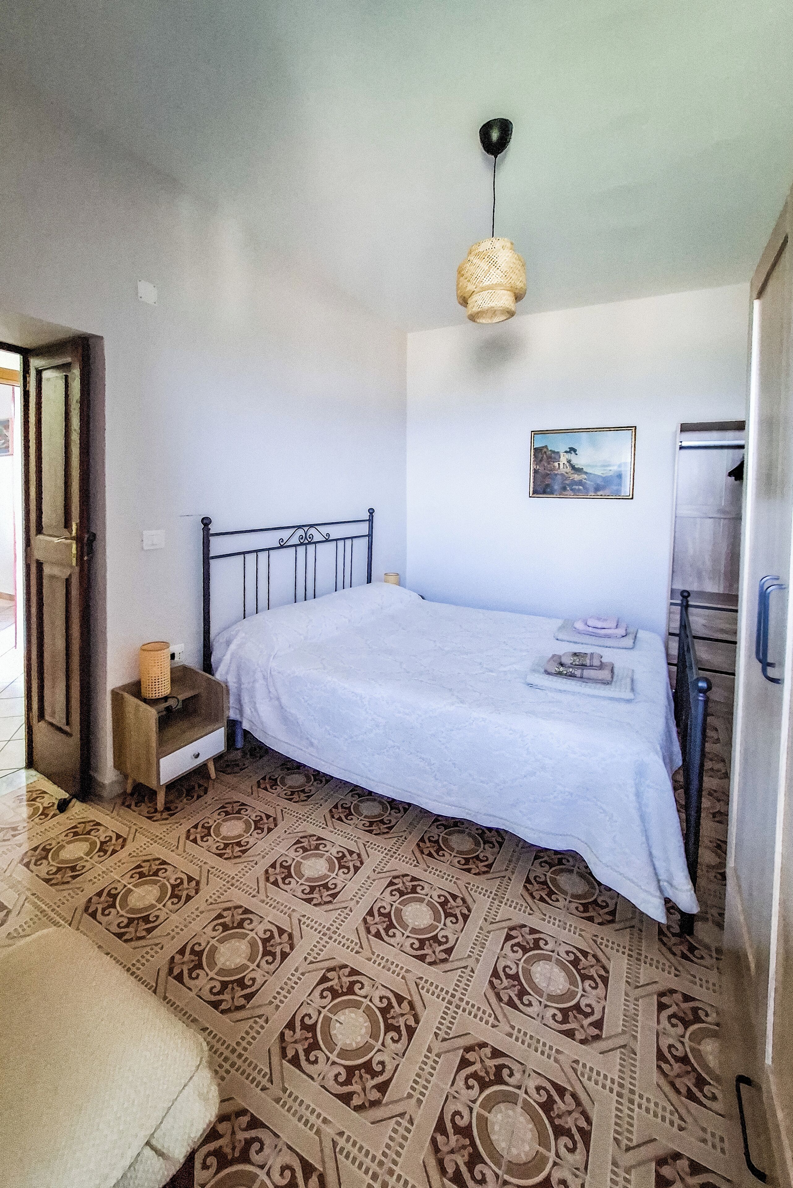 A bedroom with a bed, a nightstand, a hanging light, a painting, and a patterned floor.