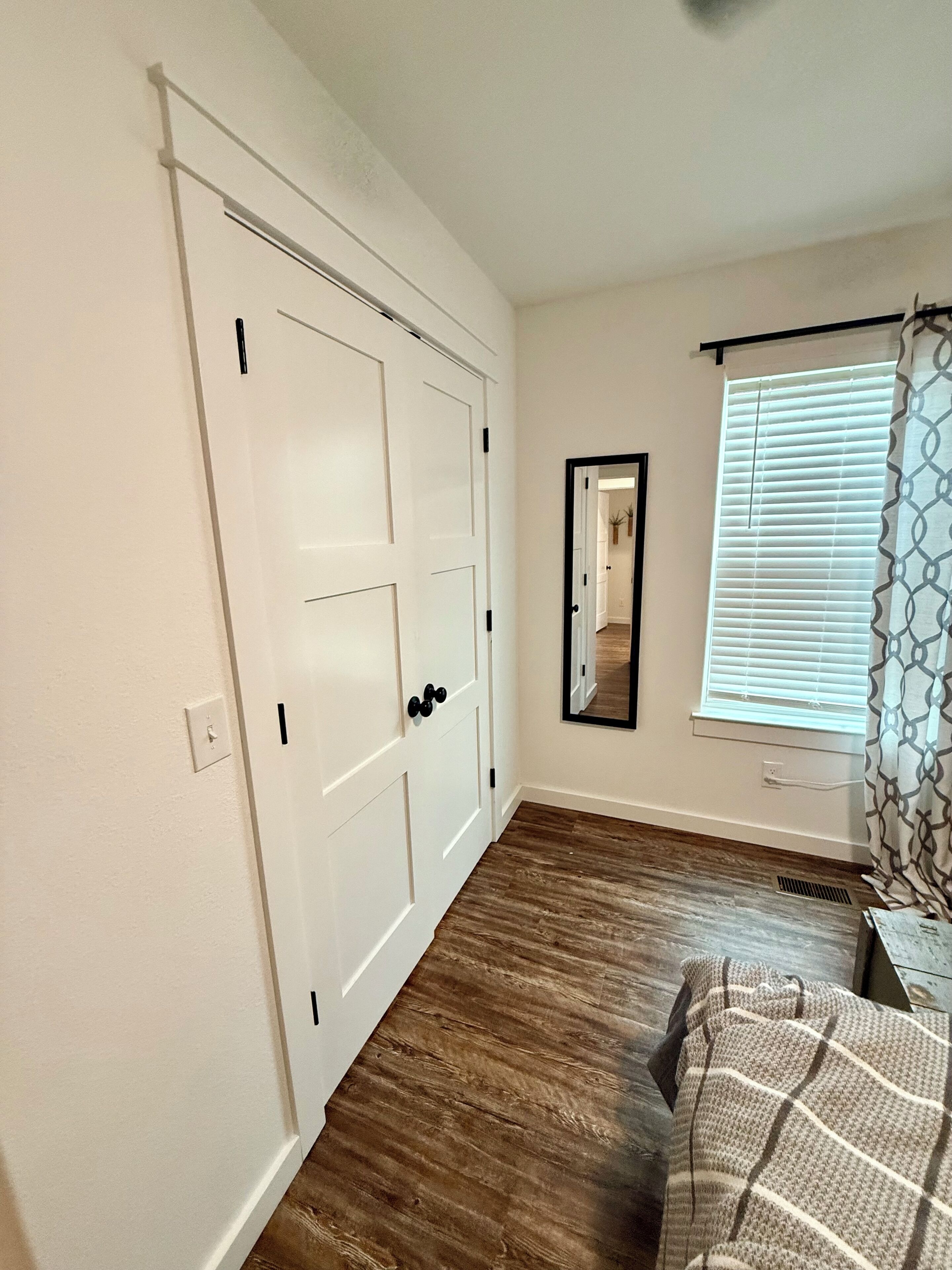 A room with a large white closet, a mirror, a window with blinds, and a wooden floor.