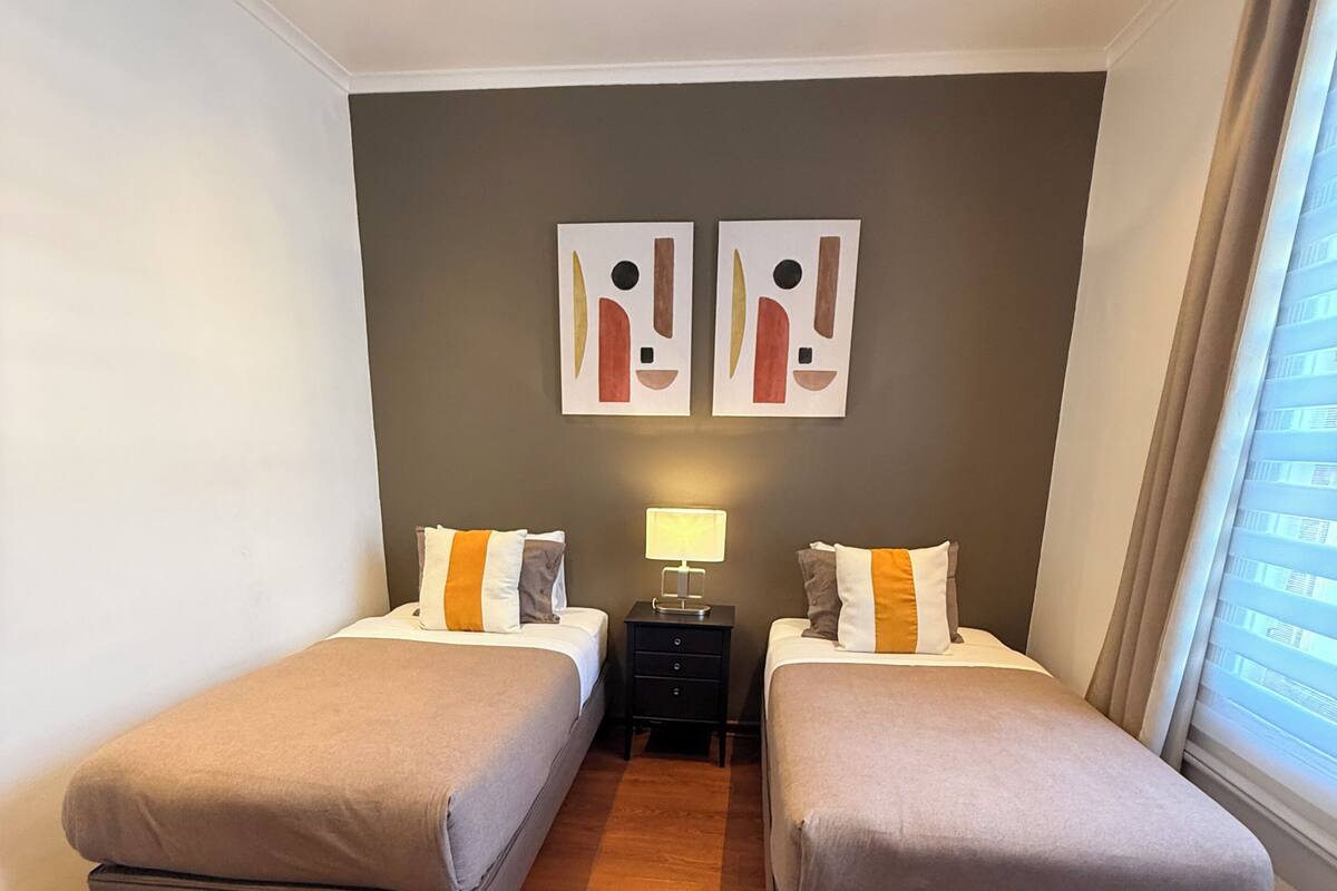 A room with two beds, a nightstand with a lamp, and abstract wall art.