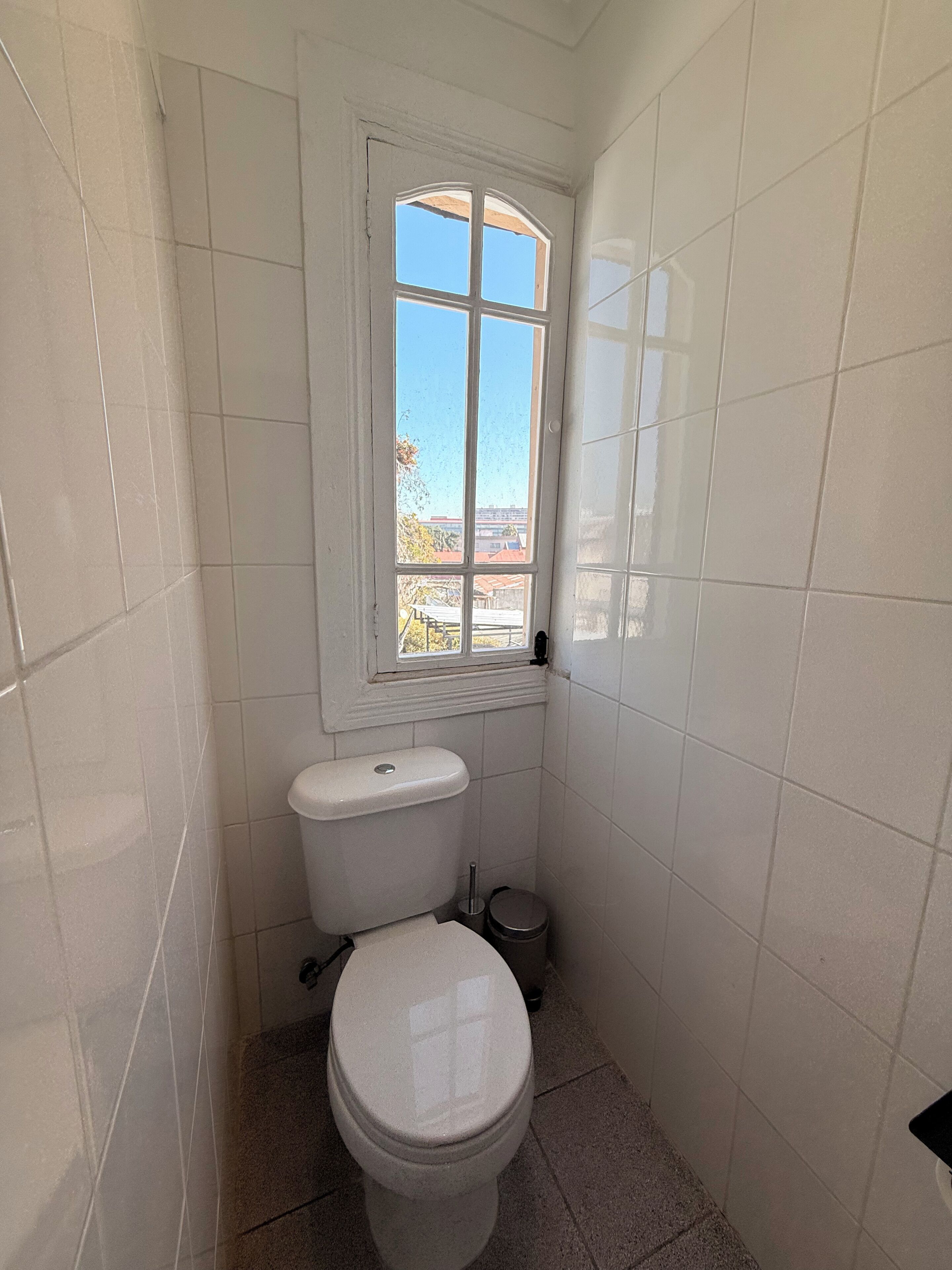 Un baño pequeño con azulejos blancos, una ventana, un inodoro y un cubo de basura.