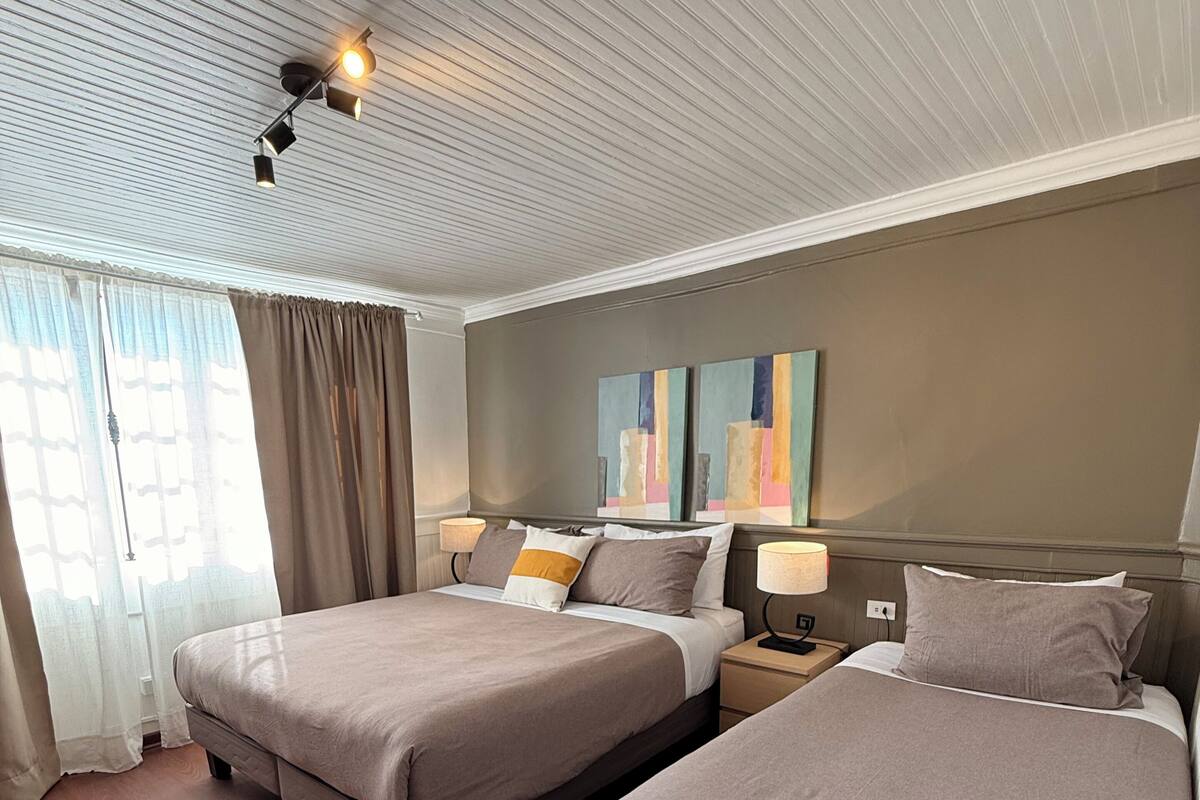 A hotel room with two beds, a ceiling light fixture, a nightstand, and a window with curtains.