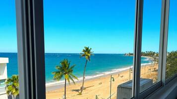 On peut admirer la plage depuis une fenêtre donnant sur une étendue de sable, bordée de palmiers, sous un ciel bleu et dégagé.