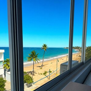 Vista de una playa desde una ventana, con arena, palmeras y un cielo azul despejado.