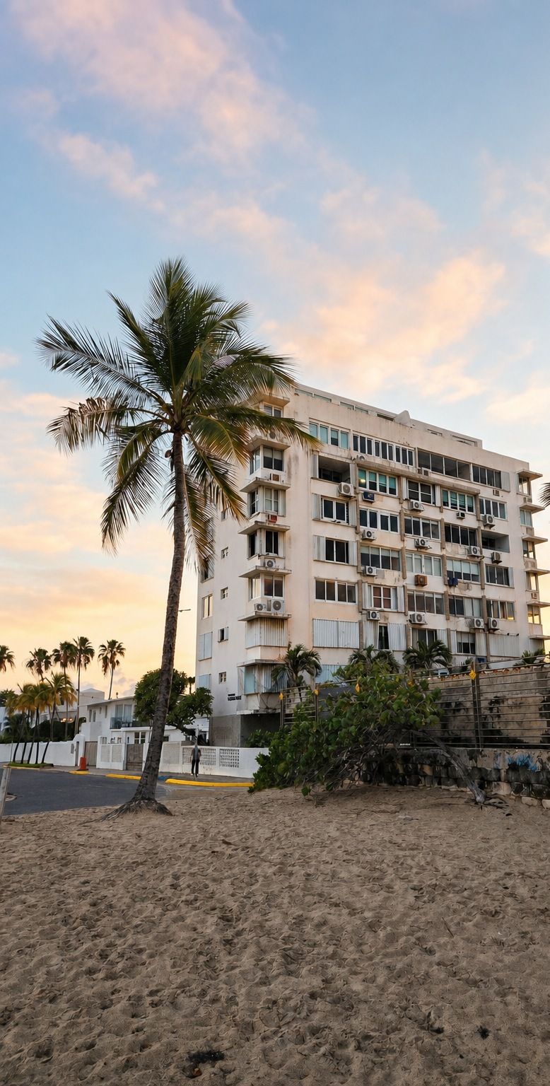 A multi-story building with balconies, a palm tree, and a sandy foreground.