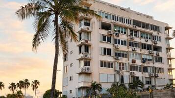 A multi-story building with balconies, a palm tree, and a sandy foreground.