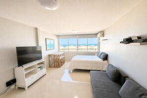 Un dormitorio moderno con una televisión, una cama, un sofá, una mesita y una ventana con vistas al mar.