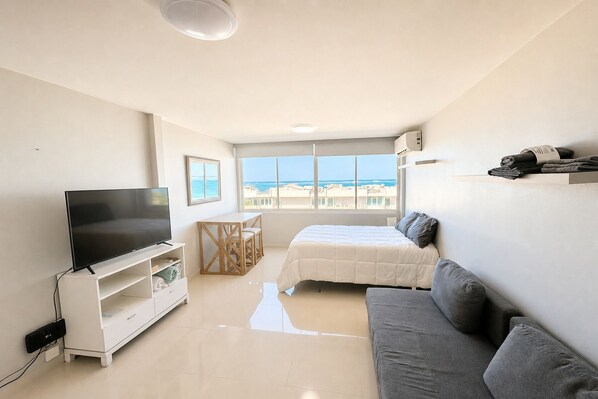 A modern bedroom with a TV, a bed, a sofa, a small table, and a window with a view of the sea.