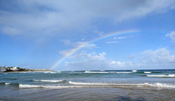 A beach with waves, a rainbow, and a coastal town in the background.