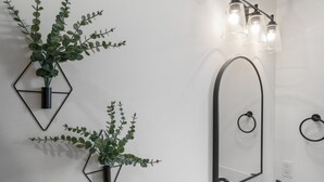 Une salle de bain avec un miroir aux motifs géométriques, un luminaire fixé au mur et deux plantes en pot.