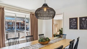 Ein Essbereich mit einem langen Holztisch, schwarzen Stühlen, einer gewebten Pendelleuchte und Blick auf ein Gebäude durch große, mit Vorhängen versehene Fenster.