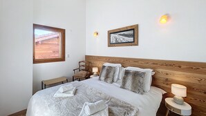 Un dormitorio con cabecera de madera, cama con sábanas blancas y varias almohadas, una mesita de noche con lámpara, una ventana con vista a una estructura de madera y dos cuadros en la pared.