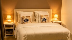 Una cama bien hecha con un edredón blanco y dos almohadas, flanqueada por dos mesitas de noche con lámparas, en una habitación con una paleta de colores neutros y cálidos.