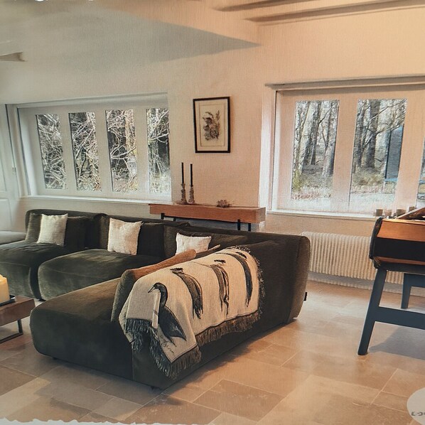 A cozy living room with a sofa and a coffee table, large windows with a view of trees, and a framed picture on the wall.
