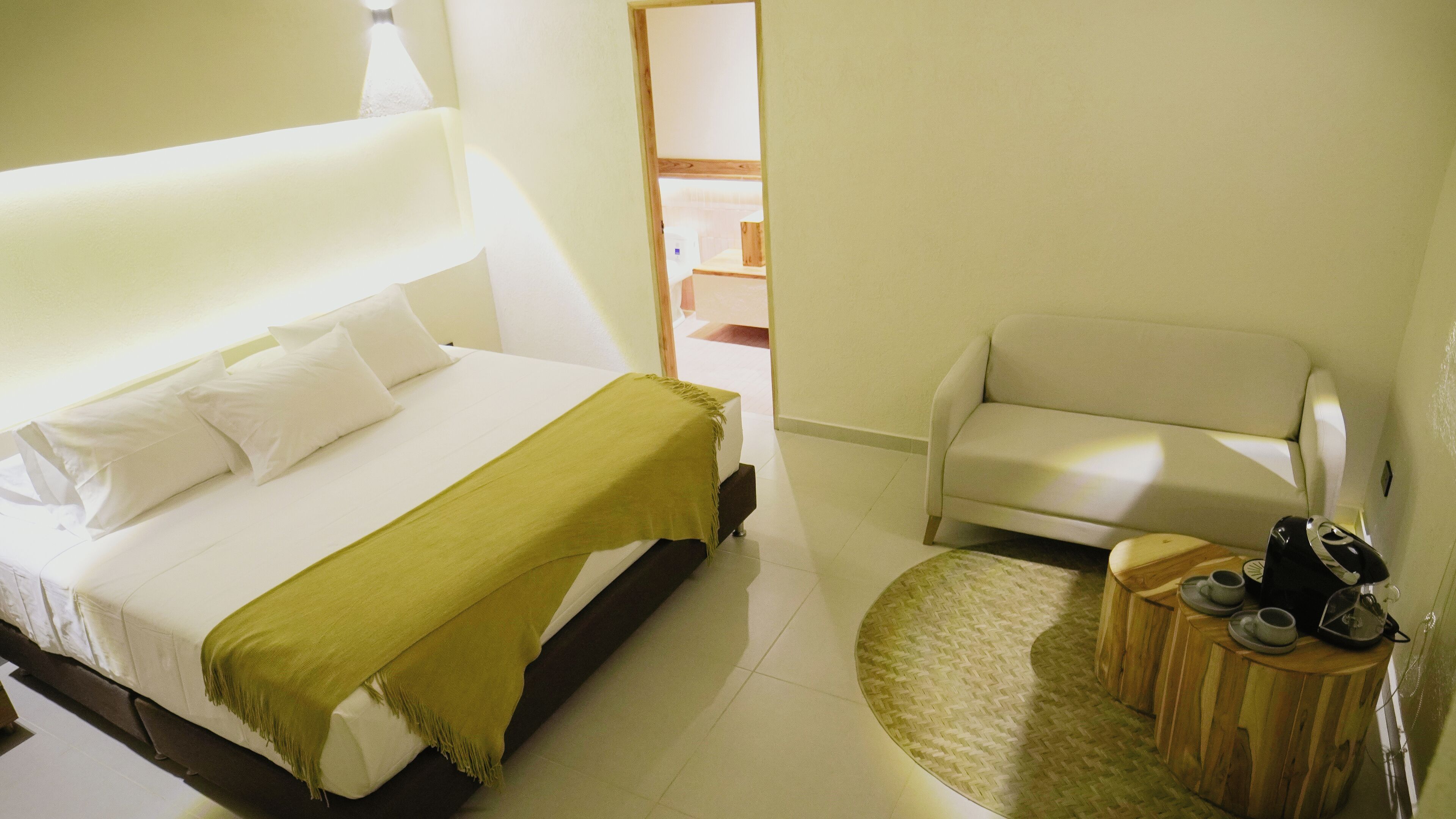 Une chambre d’hôtel avec un lit, une chaise, une petite table en bois avec des tasses et une bouilloire, et un miroir qui reflète un couloir.