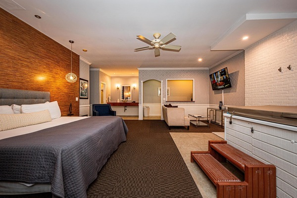 Suite features a king bed with gray quilt, a white brick hot tub with wooden steps, and a wall-mounted TV.