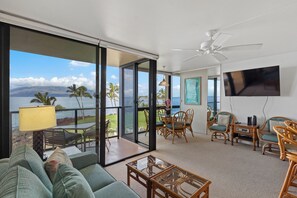 Un vaste espace de vie avec une grande porte-fenêtre coulissante donnant sur un balcon avec vue sur l’océan. On y trouve un coin salon confortable avec un canapé et des fauteuils, un téléviseur à écran plat fixé au mur et un ventilateur de plafond.