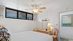 Een slaapkamer met een bed, een plafondventilator, een dressoir met spiegel, een stoel en een raam met jaloezieën.
