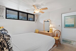 Ein Schlafzimmer mit Bett, Deckenventilator, Kommode mit Spiegel, Stuhl und Fenster mit Jalousien.