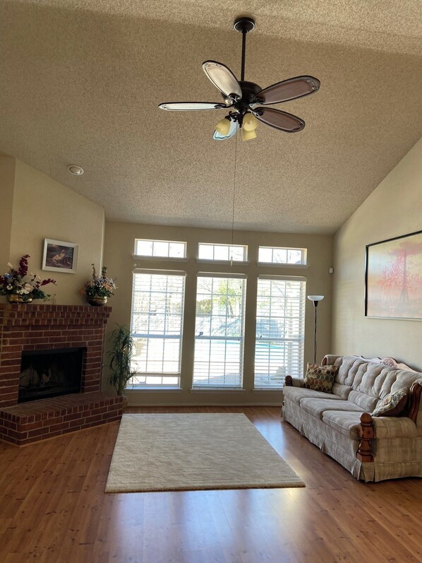 A spacious living room with a fireplace, a ceiling fan, large windows, and a cozy couch.