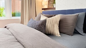 Een opgemaakt bed met een grijze deken en meerdere kussens, een raam met gordijnen, een wandlamp en behang met een geometrisch patroon.