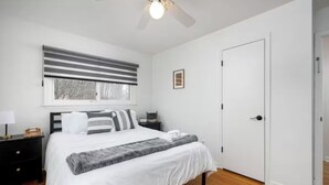 A bedroom with a bed, a nightstand, a bedside table, a window with blinds, a ceiling fan, and a door. - Walk to the Big House, Sleeps 8, 4 bedrooms, 2 full bathrooms, 3 living rooms  (Ann Arbor)