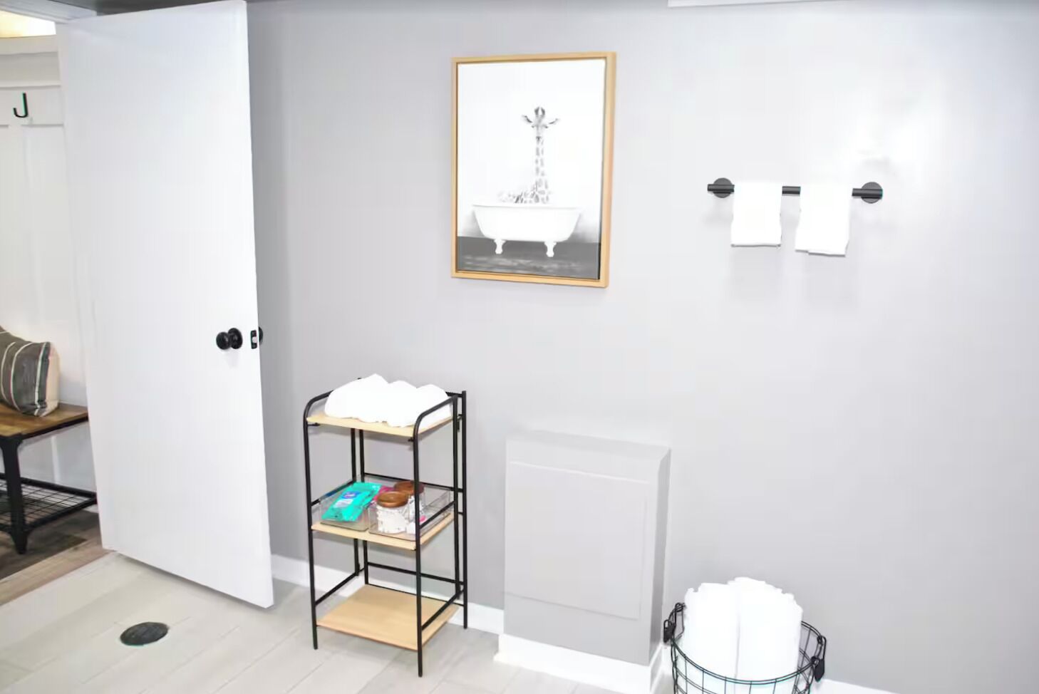 A bathroom with a white door, a framed picture of a bathtub, a towel rack, and a shelf with toiletries.