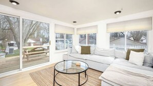 A modern living room with a glass coffee table, a white sofa, and large windows offering a view of the backyard. - Walk to the Big House, Sleeps 8, 4 bedrooms, 2 full bathrooms, 3 living rooms  (Ann Arbor)