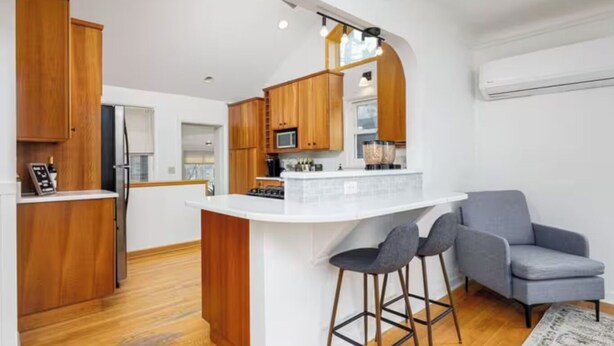 A modern kitchen with wooden cabinets, a white island with bar stools, and a grey armchair. - Walk to the Big House, Sleeps 8, 4 bedrooms, 2 full bathrooms, 3 living rooms  (Ann Arbor)
