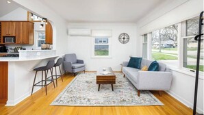 A modern kitchen with a central island, bar stools, and a grey sofa in a living area with a large window. - Walk to the Big House, Sleeps 8, 4 bedrooms, 2 full bathrooms, 3 living rooms  (Ann Arbor)