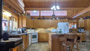 A spacious kitchen with wooden cabinets, a central island, a stove, and a dining area with chairs. - Log Home close to Hiking Trails! (Seeley Lake)