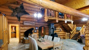 A log cabin interior with a dining area, wooden ceiling beams, a fireplace, and a bear decoration. - Log Home close to Hiking Trails! (Seeley Lake)