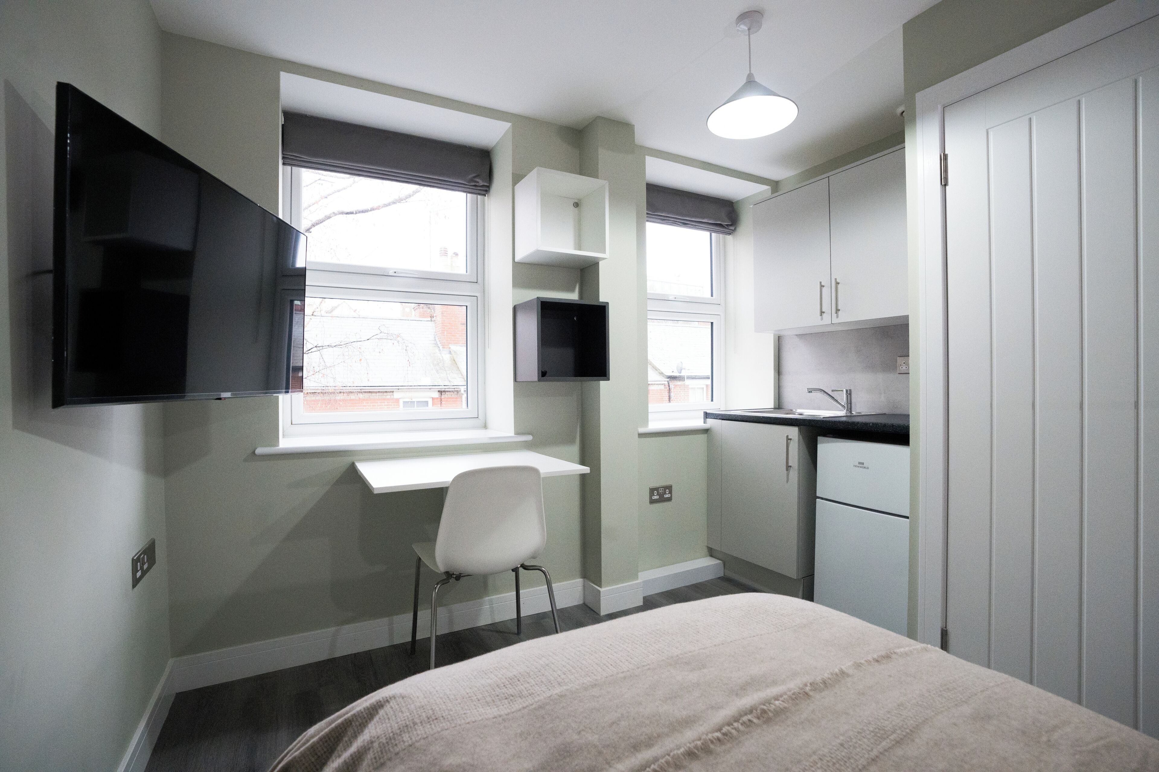 Een kleine, moderne slaapkamer met een bed, een bureau, een stoel, een aan de muur bevestigde televisie en een raam.
