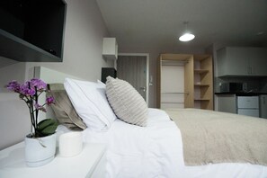 A modern hotel room with a neatly made bed, a bedside table with a plant, a white pillow, and a beige throw blanket.